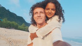 Young happy Indian woman sits on back of circling Caucasian man and screams with pleasure recording video selfie together for world travel blog located on sandy beach near ocean. Leisure, lifestyle