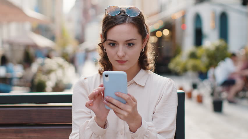 Oh my God, Wow. Amazed woman use mobile smartphone celebrating win, good message news, lottery jackpot victory, giveaway online. Happy girl sits on bench on city sunny street outdoors. Lifestyles Royalty-Free Stock Footage #1106232325