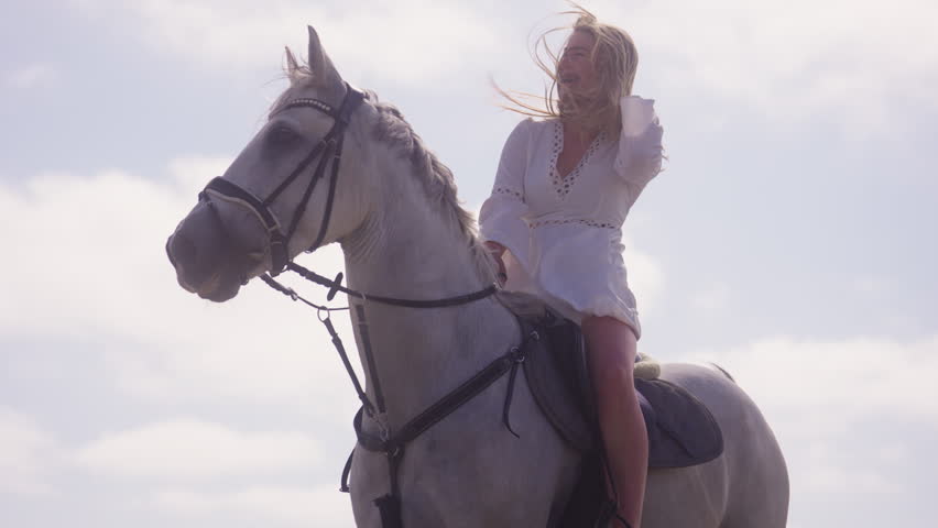 Low-angle shot of a girl wearing a white dress on a horse, fixing her hair during a windy and sunny day