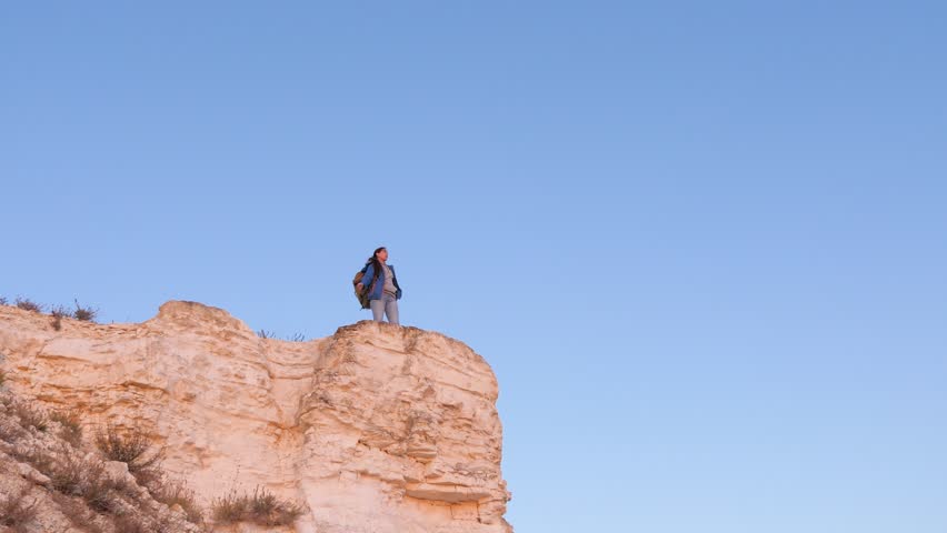Girl hiker on top of cliff against background of blue sky. Happy woman traveler approaches edge of mountain with raised hands and enjoys beautiful landscape. Free woman tourist stands on edge of cliff