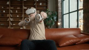 Blonde bearded man sitting on sofa at home and making hands gestures playing VR video games in slow motion 4K. Cinematic portrait of young hipster man using Virtual Reality headset with joysticks - Powered by Shutterstock - Get 15% off with code: PIKWIZARD15