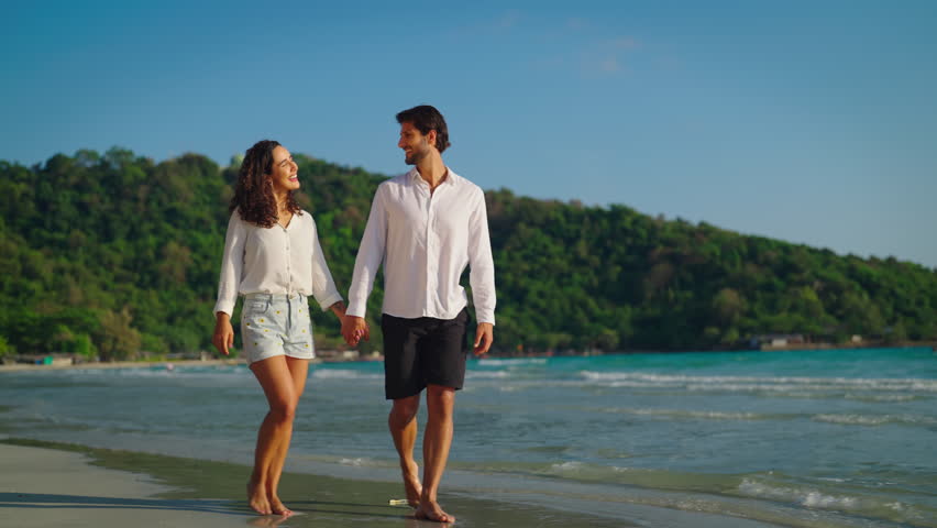 Young couple takes a leisurely stroll along the beach, their smiles beaming as they chat and laugh together, enjoying each other's company in the peaceful setting. Slow motion shot. Royalty-Free Stock Footage #1106254727