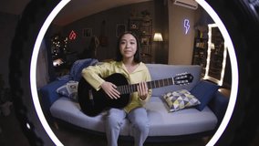 Asian girl filming a guitar lesson while sitting on sofa in front of a lamp, filming on camera through ring lamp. Talented enthusiastic blogger filming video for vlog. Concept of virtual life