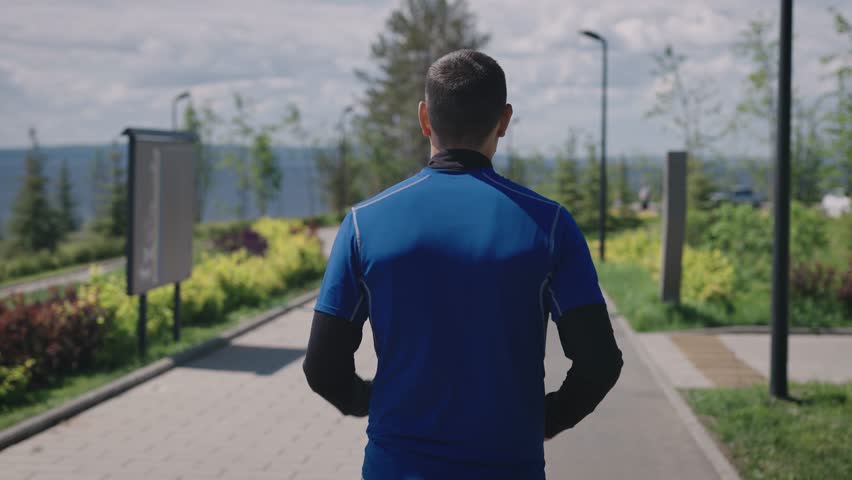 View from the back. A man in sportswear runs through a landscaped city park. Public urban spaces for sports and recreation. Royalty-Free Stock Footage #1106263039