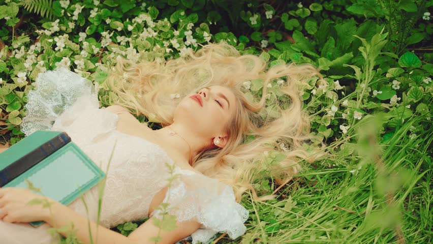 Art fairy tale sleeping beauty face girl princess blue eyes closed. Fantasy woman lies on green grass summer nature bed flowers. white medieval vintage dress. Blonde hair sexy lady read novel book. 4k