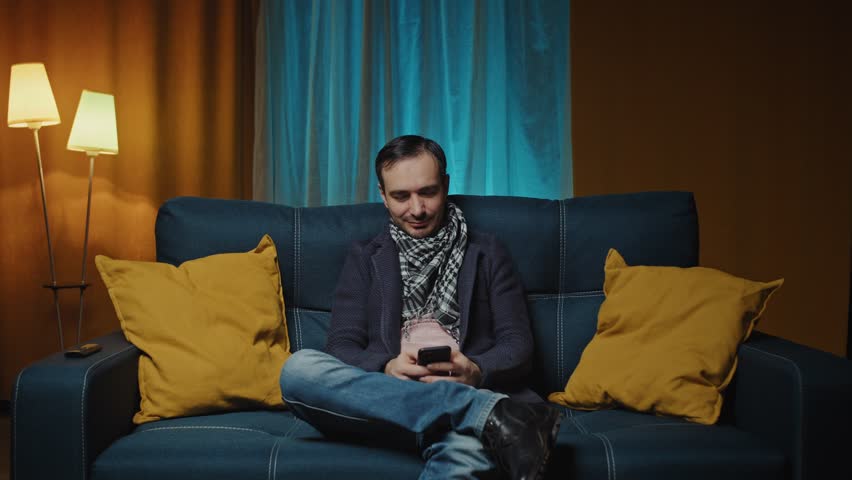 Young caucasian man relaxing on couch using smart phone,typing message in modern house.