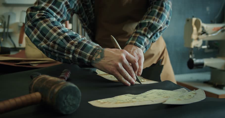 Young male bootmaker cutting out leather pieces on table at the workshop. Male tailor passing around patterns on leather pieces, preparing blanks for making shoes. Shoes making process