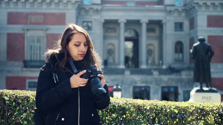 Pensive young Latin woman in warm clothes with professional photo camera looking at camera while standing near sculpture at Prado Museum in Madrid, Spain