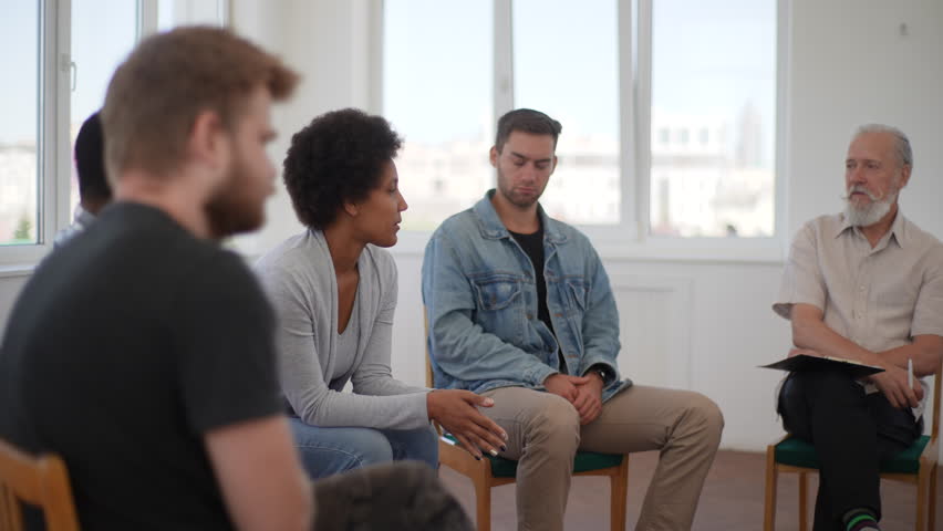 Side view of African young woman sharing sad story with multiethnic people and senior male psychologist at group therapy session. Black female dealing with addiction, talking to patients in circle.