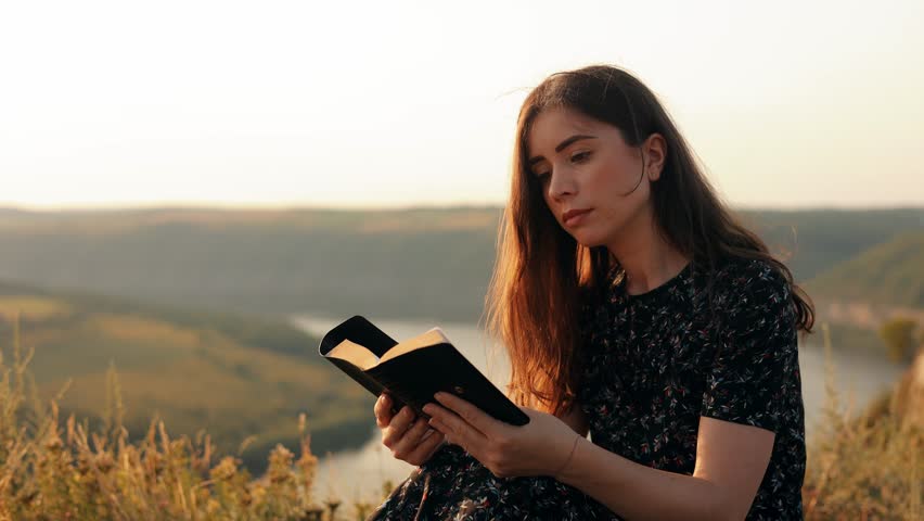 A long-haired girl reads a book in the sun. A young woman reads the Bible outdoors. A woman holds a Bible in her hands and studies the word of God on top of a mountain.  Royalty-Free Stock Footage #1106280327