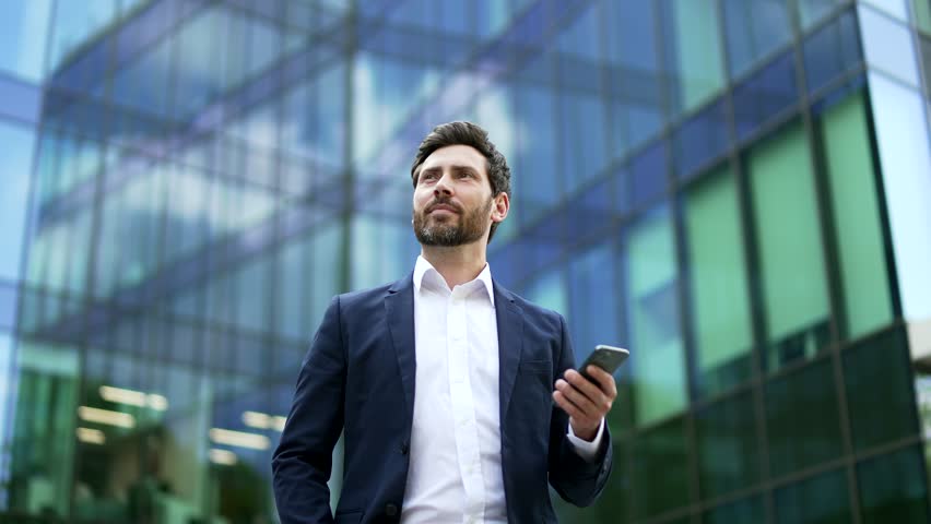 Confident mature bearded businessman in suit walking near modern glass business office building holding smartphone in hands. Man entrepreneur, investor, seo or manager using a mobile phone outside Royalty-Free Stock Footage #1106282353