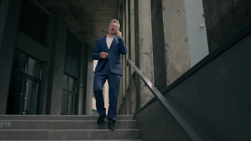 Caucasian mature old man businessman senior business employer walking outdoors on stairs near office building talking phone check time looking at wrist watch late rush hurry worried running lateness