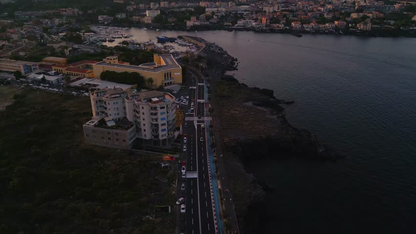 Aerial view of a seafront road at cliff beach on the volcanic rock coast of the Mediterranean sea in Catania city in Sicily, Italy at sunset with Etna volcano in the background. Royalty-Free Stock Footage #1106285795