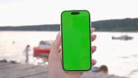 Hand holding vertical cell mobile phone with green chromakey screen in front of sea waves. Template mock up video footage. Nature background