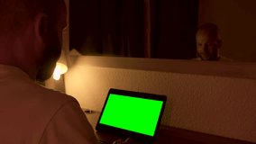 Man sitting at his desk, works on a laptop with green chroma key screen. Media. Man uses portable computer for work at home at night in front of mirror.