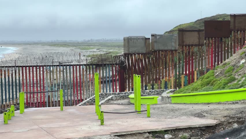 Camera pans along the border wall that separates the United States and Mexico. On the other side, in the distance, we see san Diego. Filmed on an overcast day in Playas de Tijuana (Mexico). | Shutterstock HD Video #1106291379