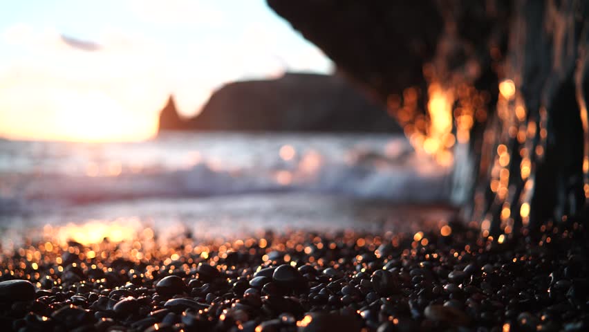 Sea sunset waves with foam. Waves run on pebbles and rock from volcanic basalt. Water breaks into splashes and white foam. Against the background of the cape and the sunset sky. Royalty-Free Stock Footage #1106292321