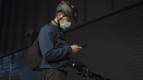Delivery man on bicycle wearing helmet and protective medical mask checks directions on mobile phone at night in city. Delivery of goods from online stores during coronavirus pandemic. Bike courier. - Powered by Shutterstock - Get 15% off with code: PIKWIZARD15
