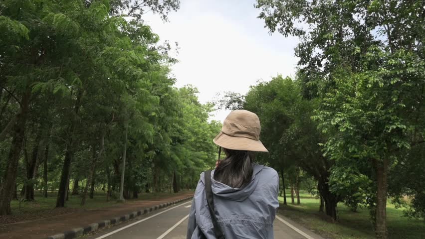Rear view of teen girl wear hat walking on the road in nature public park during summer. Outdoor pursuit. Travel destination. Environmental exploring. Slow motion.