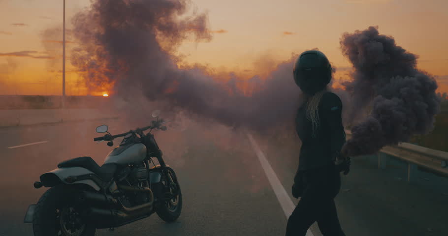 Woman with helmet and motorcycle jacket standing near her modern motorbike, smoking signal flare in her hand. Fireworks burning, highway, sunset Royalty-Free Stock Footage #1106312943