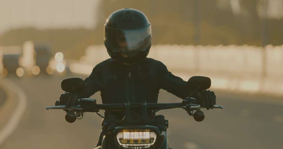 Woman biker in helmet riding modern classic motorbike on highway. Motorcyclist driving his motorcycle on asphalt route at sunset sun lighting, adventure and lifestyle, close up
