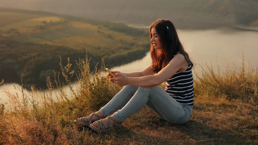 Young smiling woman listening to music in headphones while sitting on top of hill outdoors on mountain with incredible view, enjoying her favorite song and nature while walking or traveling