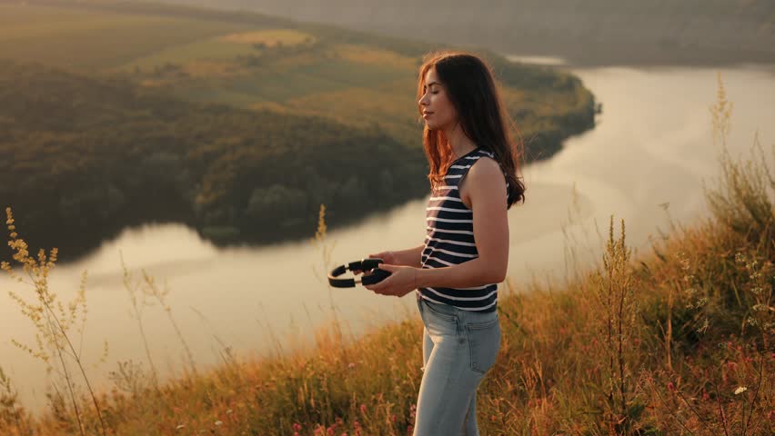 
A young girl on the background of the nature of the river and mountains enjoys music in headphones at sunset, sings along and dances cheerfully and amusingly looking at the camera