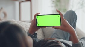 Closeup - Happy young woman holding mobile smartphone with green screen while resting on the sofa in living room at home. Watching movies on the phone.