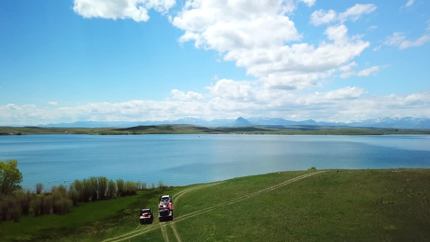 Aerial Panning Shot Of Vehicles On Road Near Lake Against Cloudy Sky, Drone Flying Over People And Dogs On Green Plants - Billings, Montana