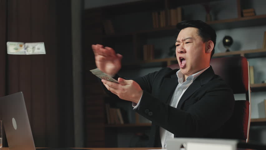 Angle view of prosperous company ceo having fun in own workplace and scattering dollar bills. Thrilled asian man wearing official stylish suit boasting wealthy life. Royalty-Free Stock Footage #1106324921
