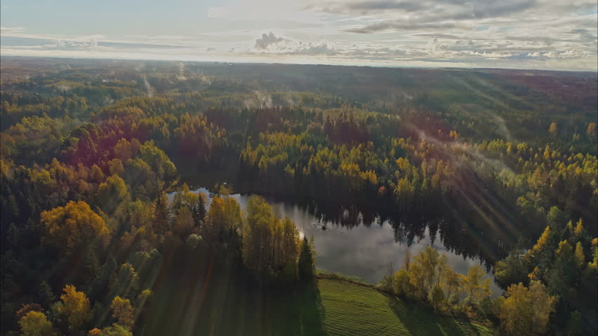 Outstanding aerial view of a countryside full of mature colorful trees, lake and a small grassy area with small fleeting clouds.