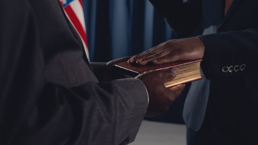 Close-up tilting shot of solemn bearded middle-aged black male politician in suit, tie and glasses taking oath on holy Bible or Quran, with raised hand, during official ceremony in front of USA flag Royalty-Free Stock Footage #1106328081