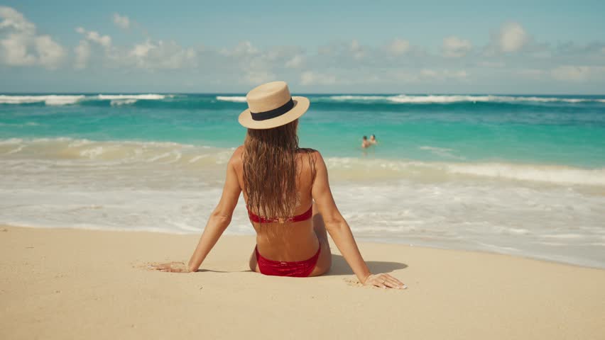 Blonde woman in red bikini straw hat sitting on ocean beach in tropical paradise island with turquoise water waves, white sand in sunny weather. Girl admire seascape. Beach resting, travel, tourism.