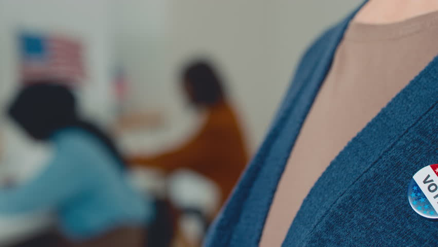 Close-up shot of election campaign vote badge on chest of unrecognizable woman in casual blue cardigan in polling station, silhouettes of women sitting in booths and USA flag in blurred background Royalty-Free Stock Footage #1106329779