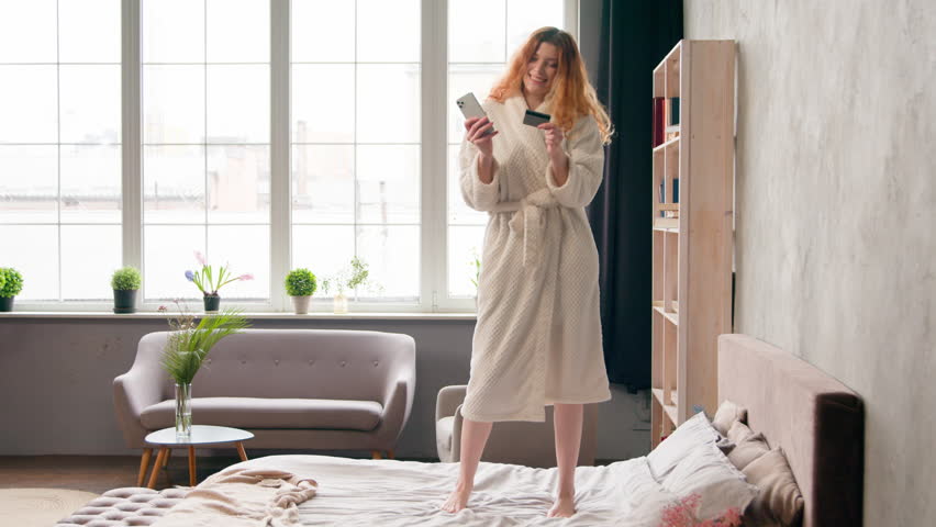 Caucasian girl buying internet online purchase booking order happy carefree woman in bathrobe shopping mobile phone and credit bank card success payment dancing celebrate discount on bed in bedroom Royalty-Free Stock Footage #1106332491