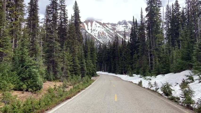 View along an empty country road lined with evergreen trees towards a snowcapped mountain and snow banks on the side of the road - North Cascades National Park, Washington, USA