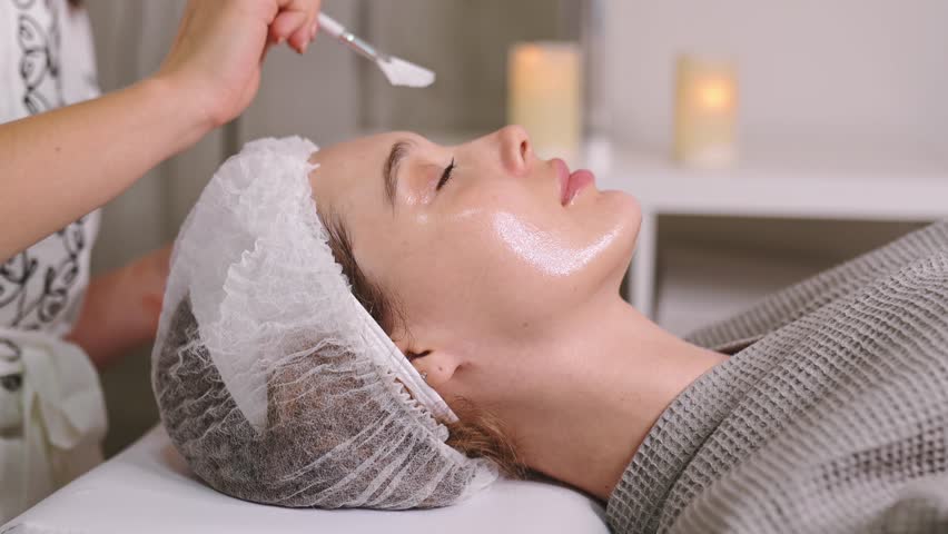 Beauty procedure. The cosmetologist cleans the face of a beautiful caucasian woman with special gel before applying a mask, peeling, scrub. Side view. Facial skin care procedures in a beauty salon Royalty-Free Stock Footage #1106335331