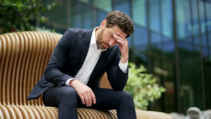 Depressed man businessman employee entrepreneur in a formal suit sitting on a bench near office building holding head. Sad male owner investor experiencing depression has problems with mental health Royalty-Free Stock Footage #1106340583