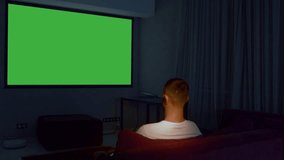 Rear view of a man sitting on sofa in living room and watching tv with green screen. Media. Guy in front of TV with green chroma key screen.