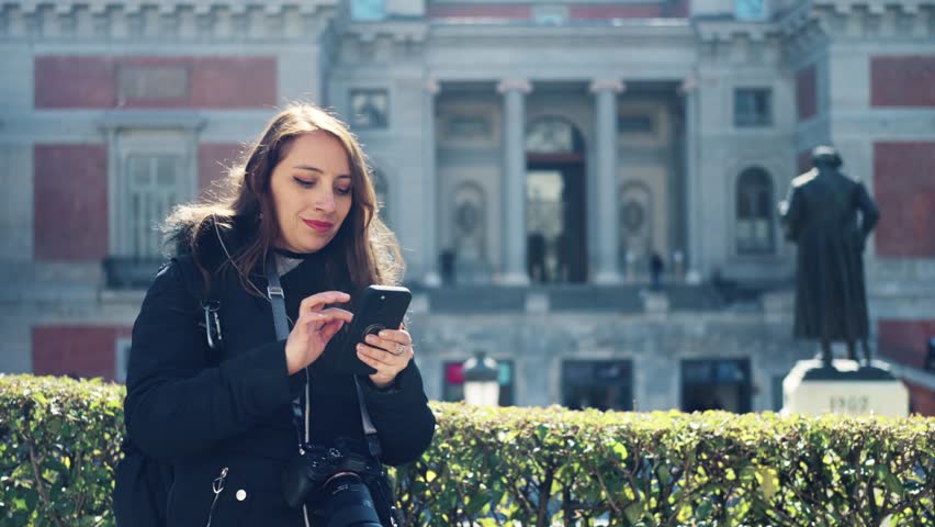 Pensive young Latin woman in warm clothes with professional photo camera using smartphone while standing near Prado Museum in Madrid, Spain