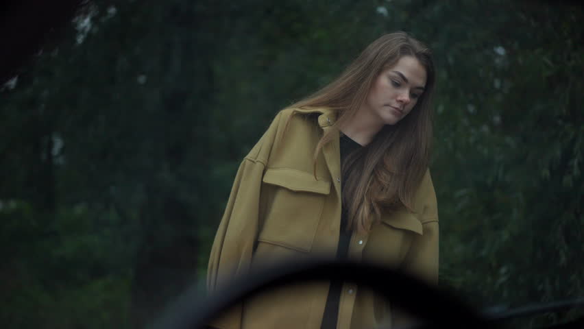 Shooting through windshield of sad unsure woman examining damaged automobile gesturing hopelessness looking at camera. Young Caucasian beautiful lady posing outdoors at broken vehicle | Shutterstock HD Video #1106354441