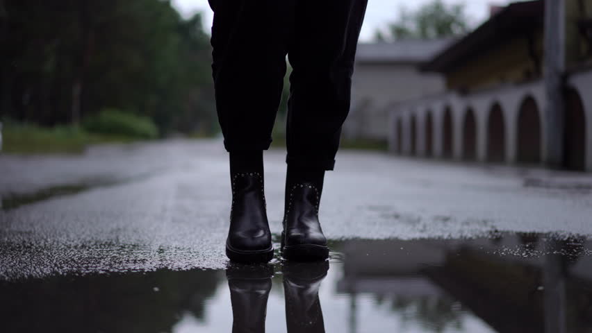 Dolly shot female Caucasian feet in black boots walking in slow motion in puddle splashing water. Front view unrecognizable young self confident woman strolling on cloudy rainy day outdoors