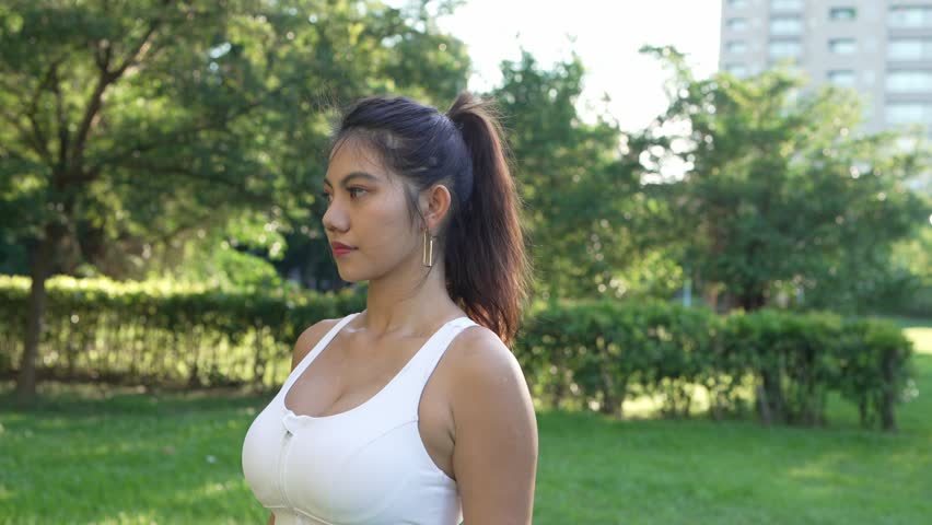 Young asian woman warming up doing jumps at a city park, preparing to jog and run outdoors on a sunny bright day.
