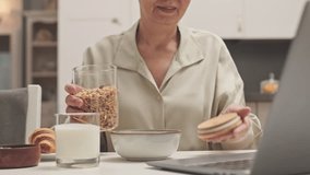 Cropped shot of adult Asian woman pouring some granola in bowl preparing healthy breakfast at home while having video chat on laptop