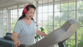 Happy asian elderly women in headphones talking and waving hands to say hello through video call on smartphone while exercising on treadmill at home. Elderly activity, Health care concept