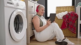 Middle age grey-haired woman watching video on touchpad waiting for washing machine at laundry room