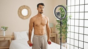 Young hispanic man having online training with dumbbells at bedroom