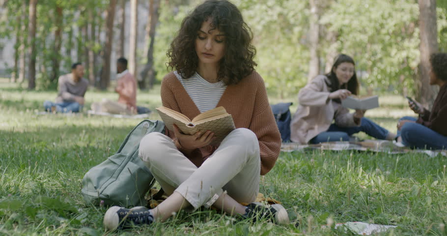 Portrait of beautiful young woman student reading book preparing for exam on lawn in park. Outdoor activity and motivated youth concept.