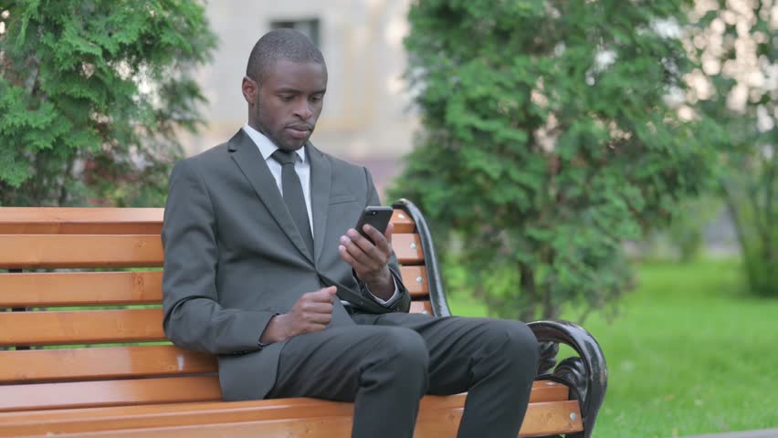 African Businessman Celebrating Success on Smartphone while Sitting Outdoor on a Bench