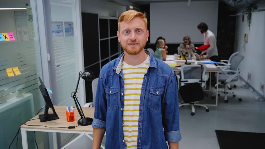 Young red-haired programmer or businessman in a modern office looking camera and smiles. Self-employed guy with his arms crossed poses for a video in the meeting room. Colleagues work in background. Royalty-Free Stock Footage #1106383643
